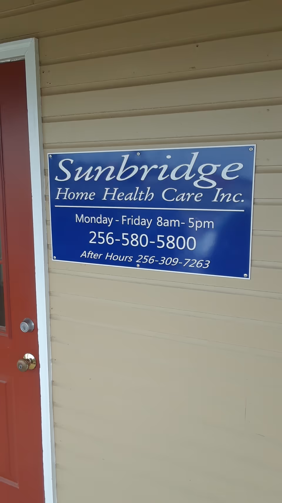 Blue Sunbridge Home Health Care Inc. sign on beige exterior wall beside a red door.