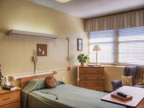 A patient room in a senior living facility with a bed, dresser, seating, and windows.