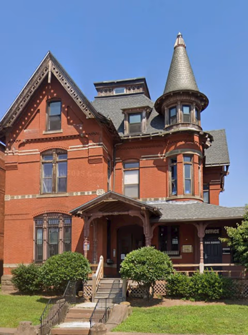 Red brick Victorian-style senior living building with turret, front porch, and greenery.