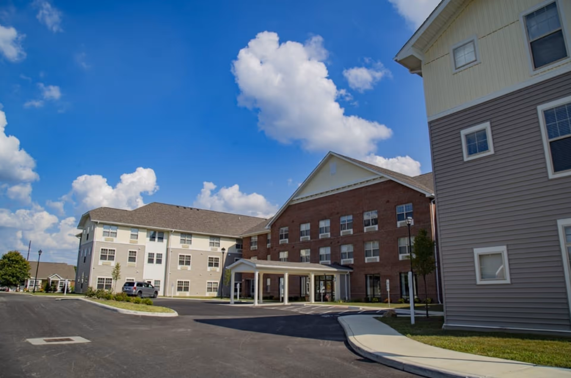 Exterior view of The Ashford at Sturbridge senior living facility showing multiple connected buildings with a mix of brick and siding facades under a blue sky with scattered clouds. There is a driveway and parking area in front with some landscaping and street lamps.