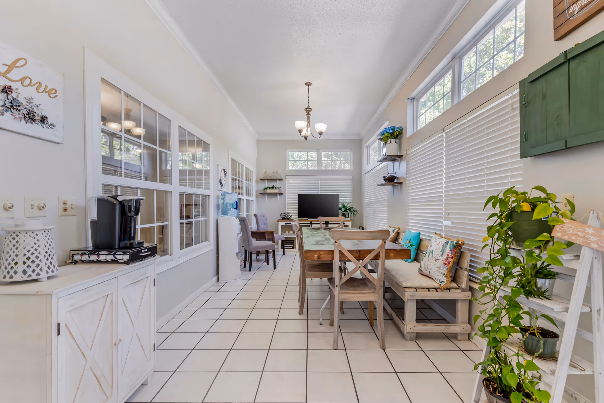 Bright communal living area with a dining table, seating, plants, and a TV.