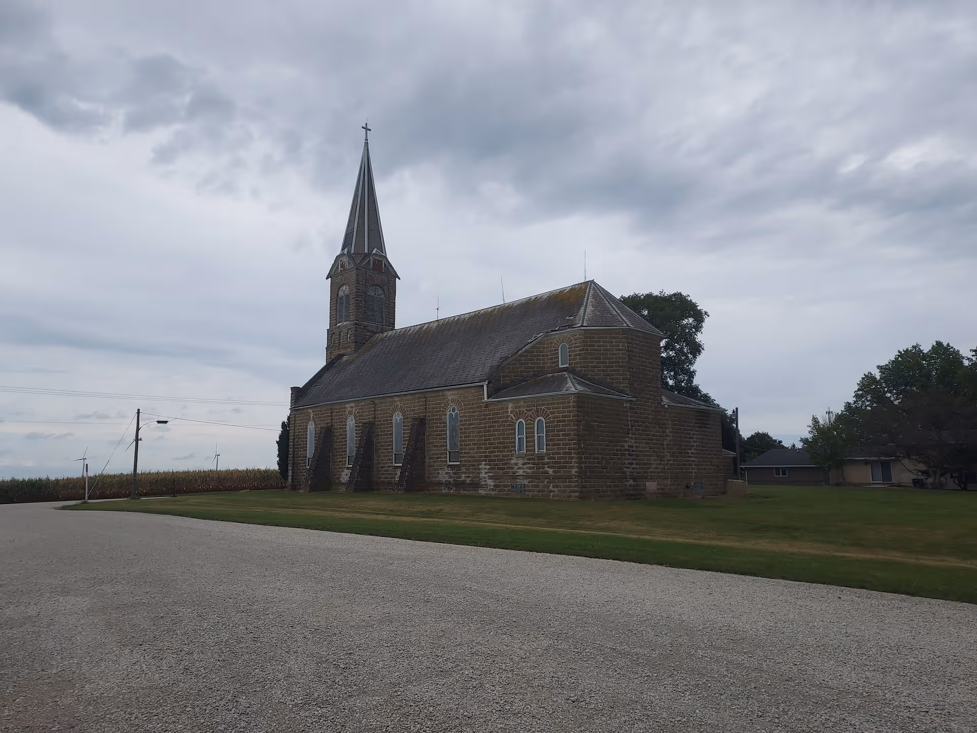 Brick church with tall steeple and cross beside a gravel road on a cloudy day.