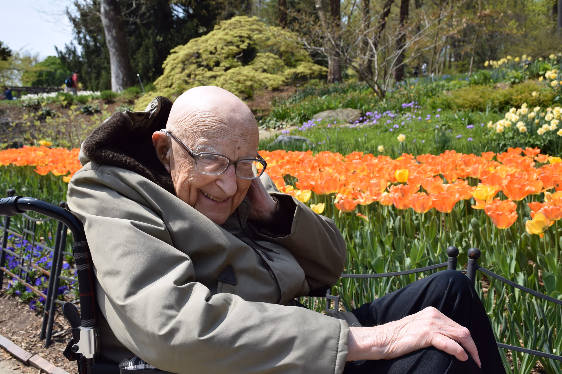 An elderly man wearing glasses and a green jacket sits in a wheelchair outdoors, smiling and touching the side of his head. Behind him is a vibrant garden filled with orange tulips, other colorful flowers, and green foliage under a clear sky.