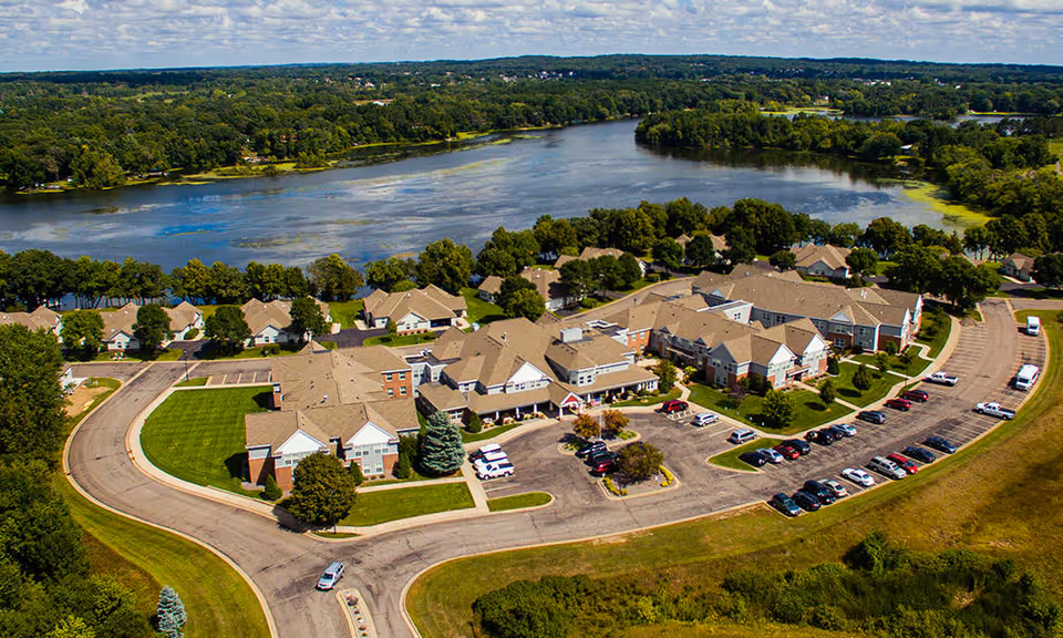 Aerial view of Guardian Angels By The Lake Senior Living facility in Elk River, showing multiple connected buildings with beige roofs surrounded by trees and greenery, a parking lot with cars, and a large lake in the background under a partly cloudy sky.