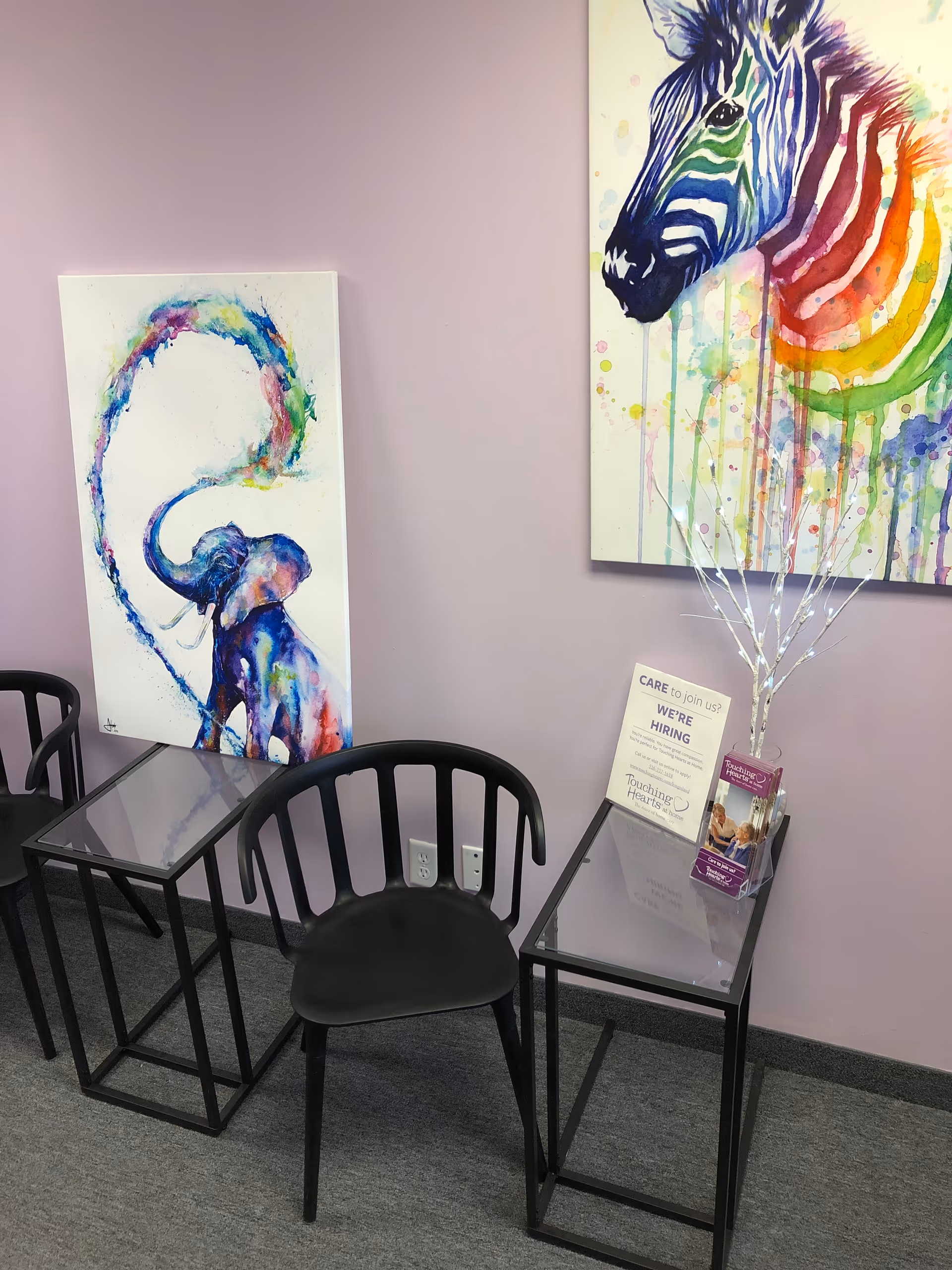 Purple-walled waiting area with black metal chairs and glass-topped tables beneath colorful artwork.