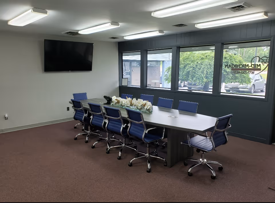 A conference room with a long gray table, blue chairs, and a wall-mounted TV.