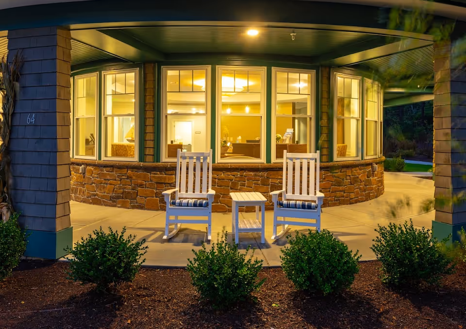 Illuminated entrance porch of a senior living facility with two white rocking chairs and a small table in front of large windows.