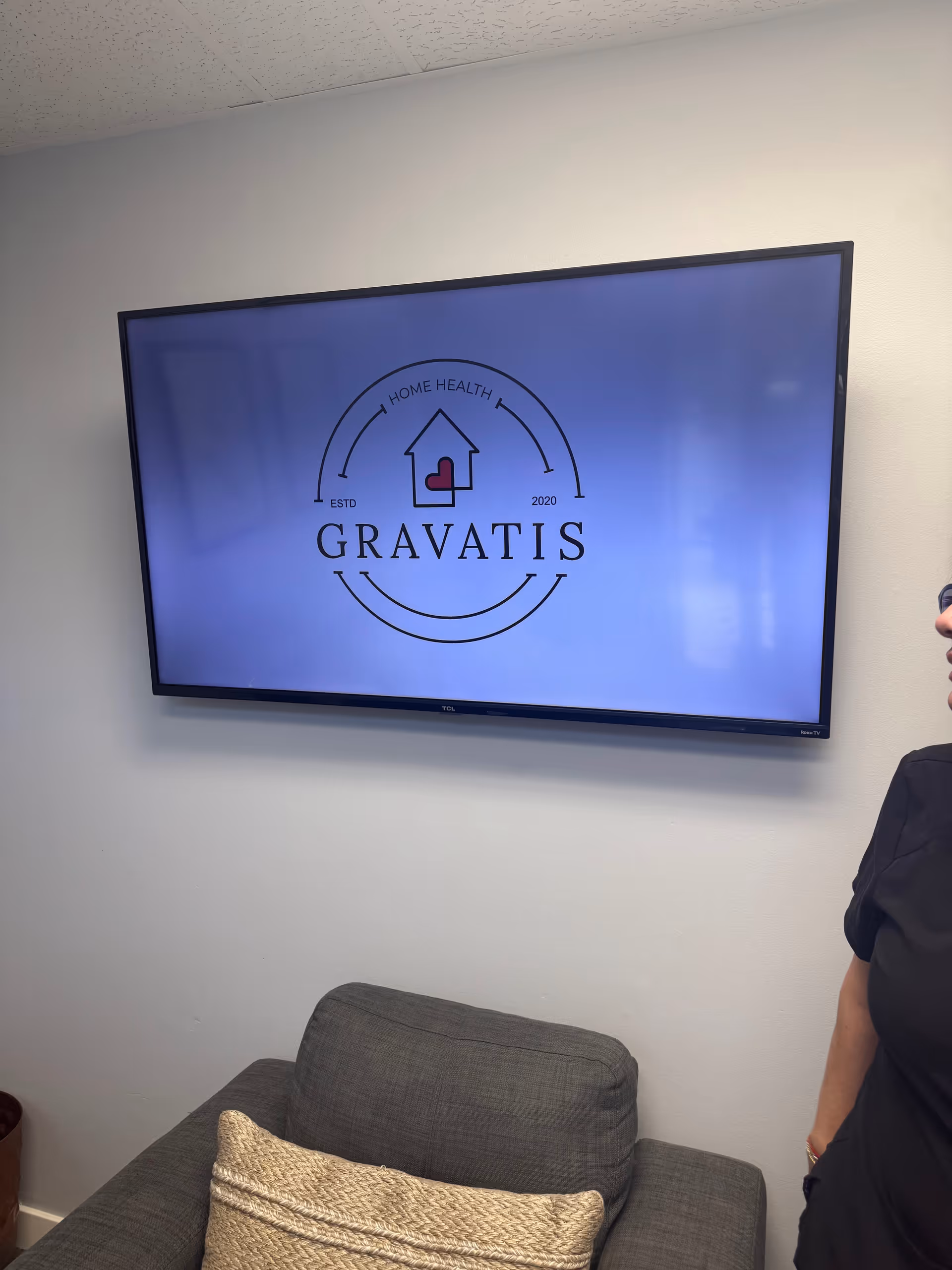 Wall-mounted TV shows Gravatis Home Health Care logo above a gray sofa.