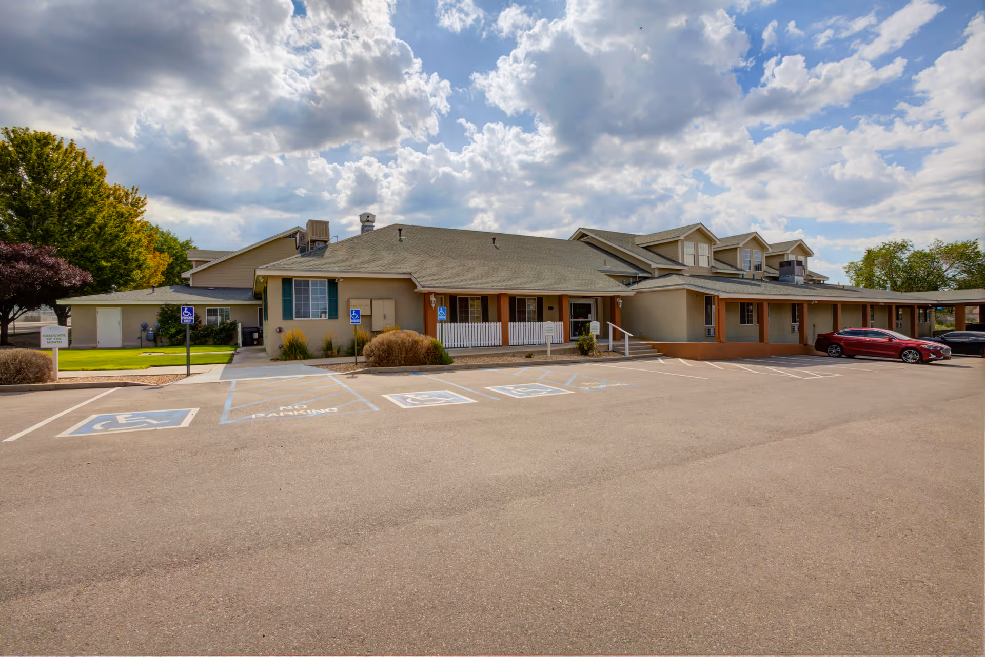Exterior view of Avista Senior Living Albuquerque building with a parking lot in front, including several handicap parking spaces. The building is single-story with a beige facade, green shutters, and a covered porch area. Trees and a partly cloudy sky are visible in the background.