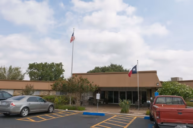 Exterior view of a single-story building with two flagpoles displaying the American and Texas flags in front. There are parked cars in the parking lot, including a silver sedan and a red pickup truck. The building has a flat roof and a covered entrance with some plants and trees around.