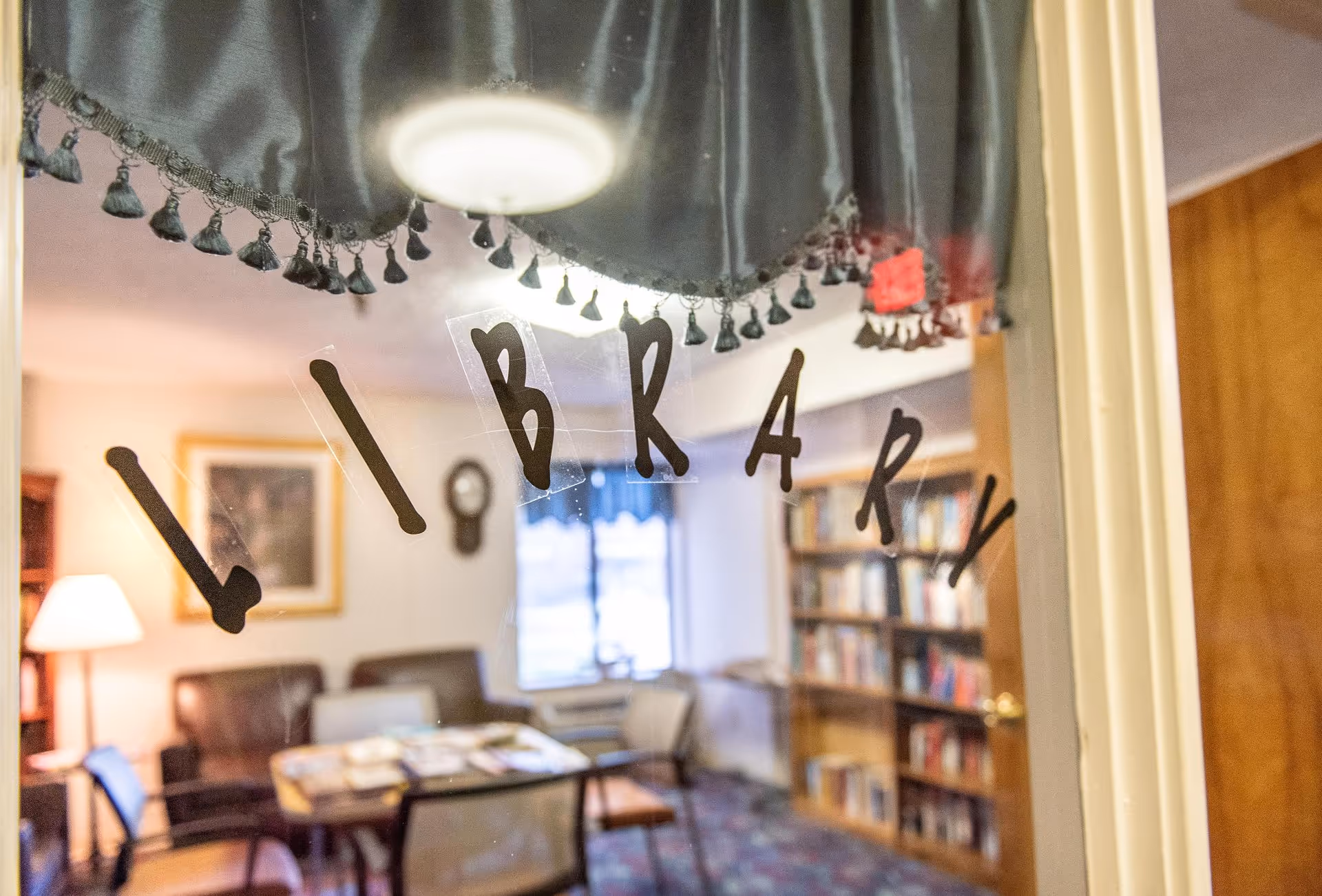 Legacy at Grande Vie library room with bookshelves, tables, chairs, and a lamp visible through glass.