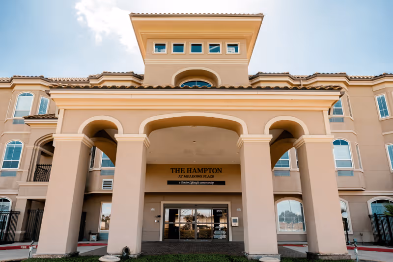Front exterior view of The Hampton at Meadows Place building with a covered entrance supported by large columns and multiple windows under a clear sky.