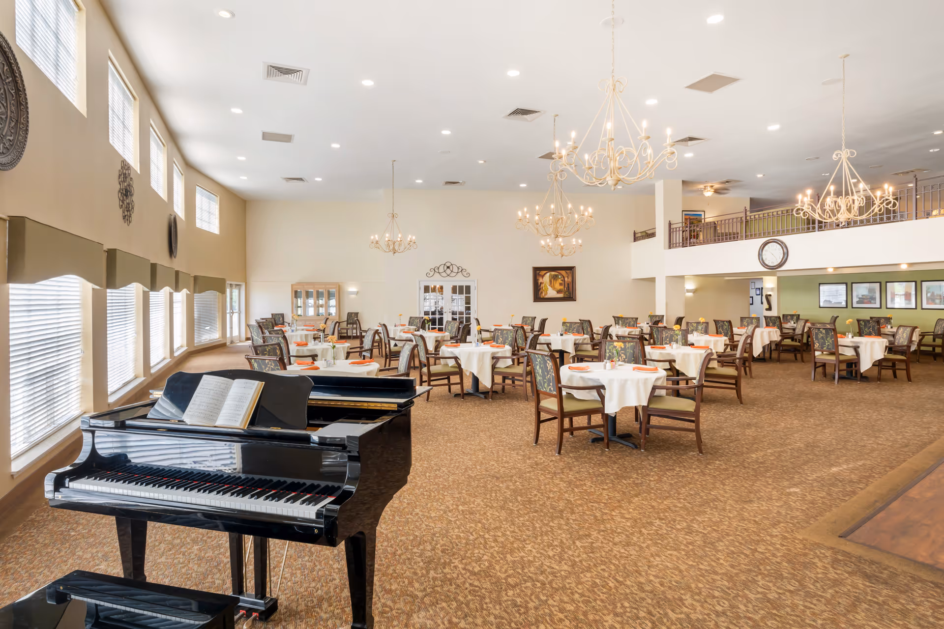 Large senior living dining hall with round tables, white tablecloths, chairs, chandeliers, and piano.