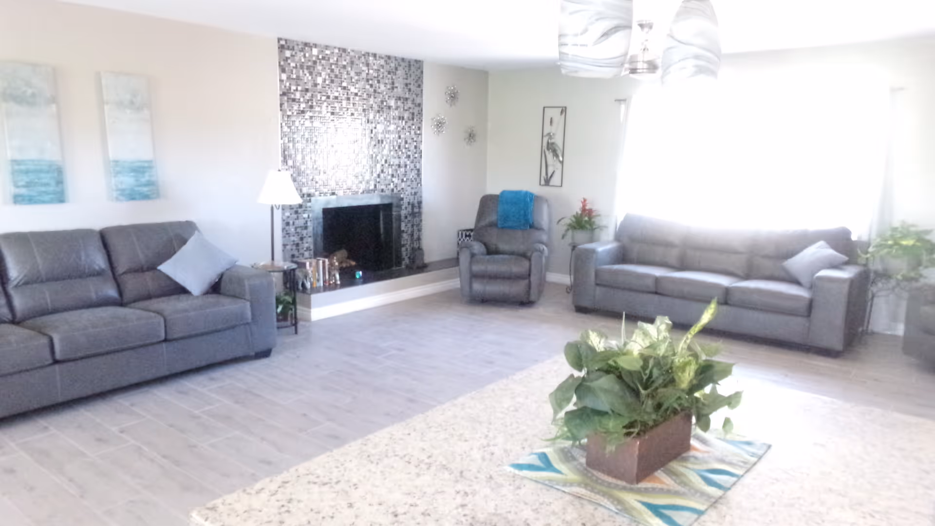 Bright living room with two gray sofas, a recliner, a mosaic-tiled fireplace, and a potted plant on a rug.