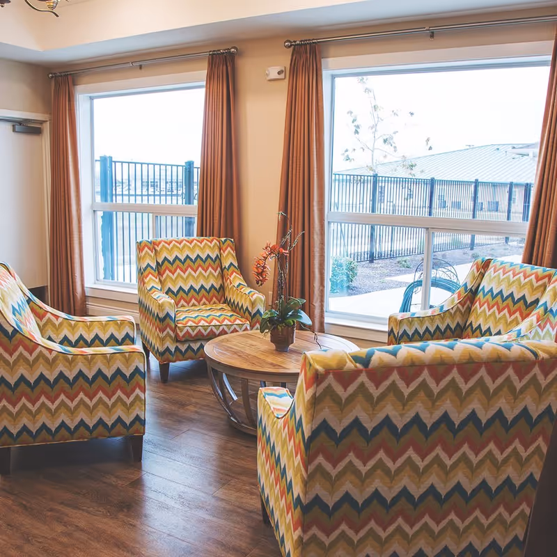 A cozy seating area with four colorful patterned armchairs arranged around a round wooden coffee table with a potted plant on it. The room has large windows with brown curtains, allowing natural light to brighten the space and showing a fenced outdoor area.