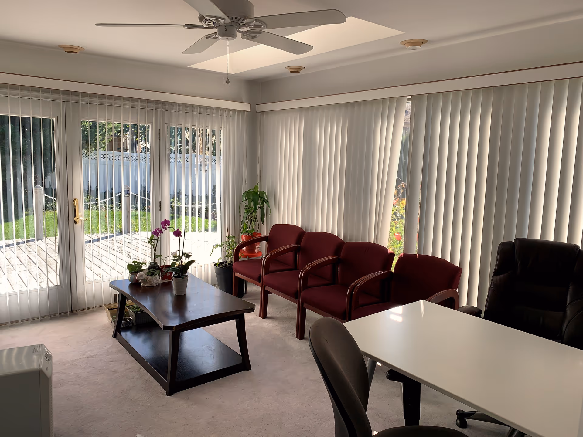 Sunlit lounge area with red chairs, a coffee table, plants, and vertical blinds.