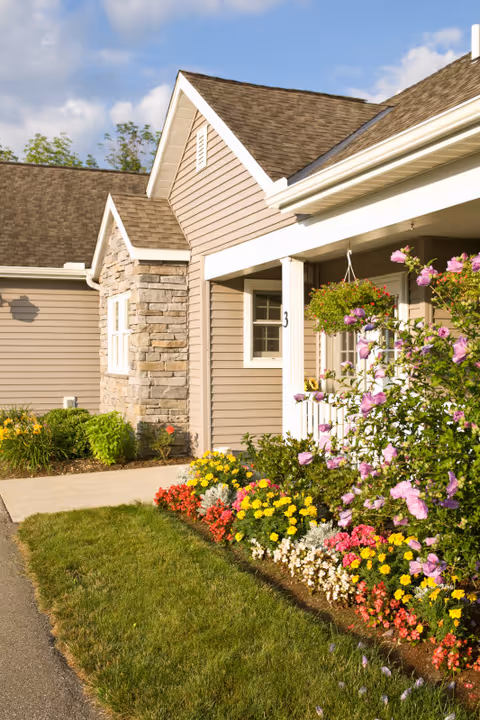 Exterior view of a senior living facility building with beige siding and stone accents, a porch with white railing, and a colorful flower garden in front under a partly cloudy sky.