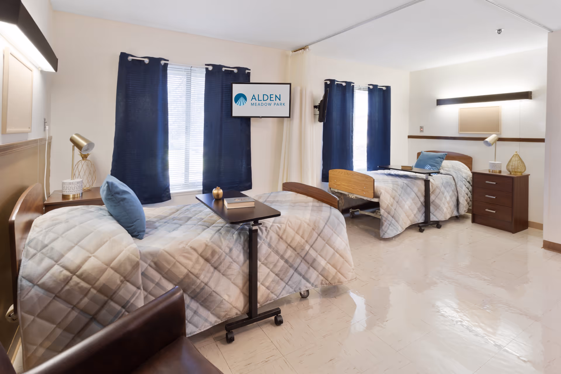 Two hospital-style beds in a shared senior living bedroom with navy curtains and a wall TV.
