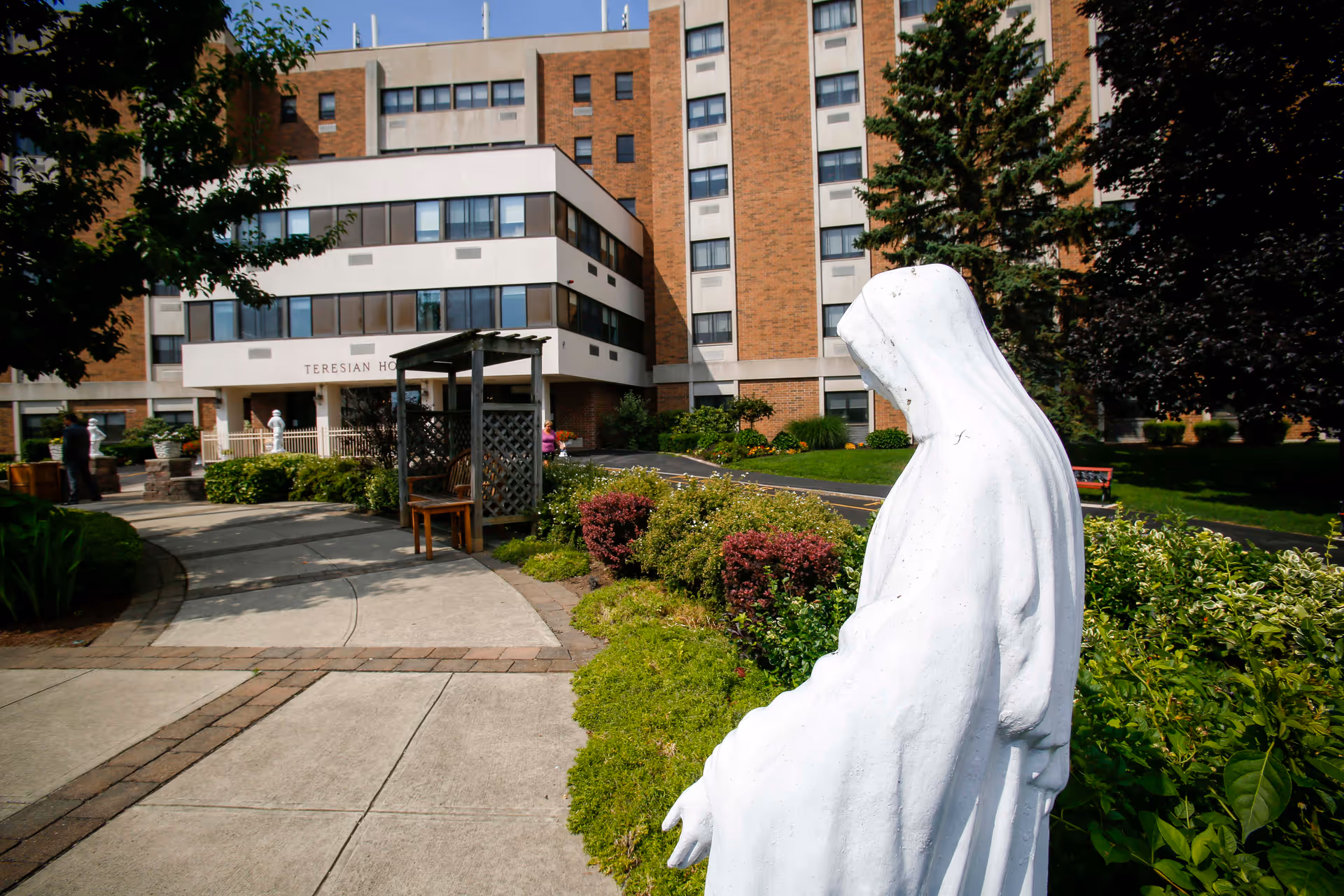 Outdoor view of Teresian House Center for the Elderly showing a multi-story brick and white building with windows. In the foreground, there is a white statue of a robed figure, likely religious, surrounded by green bushes and a paved walkway leading to the building entrance. Trees and landscaped greenery are also visible.