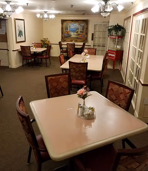 Interior view of a dining room with several square tables and cushioned chairs arranged around them. Each table has a small flower arrangement and a condiment set. The room is softly lit with ceiling lights and decorated with framed artwork on the walls. There is a popcorn machine in the corner near glass-paneled doors.