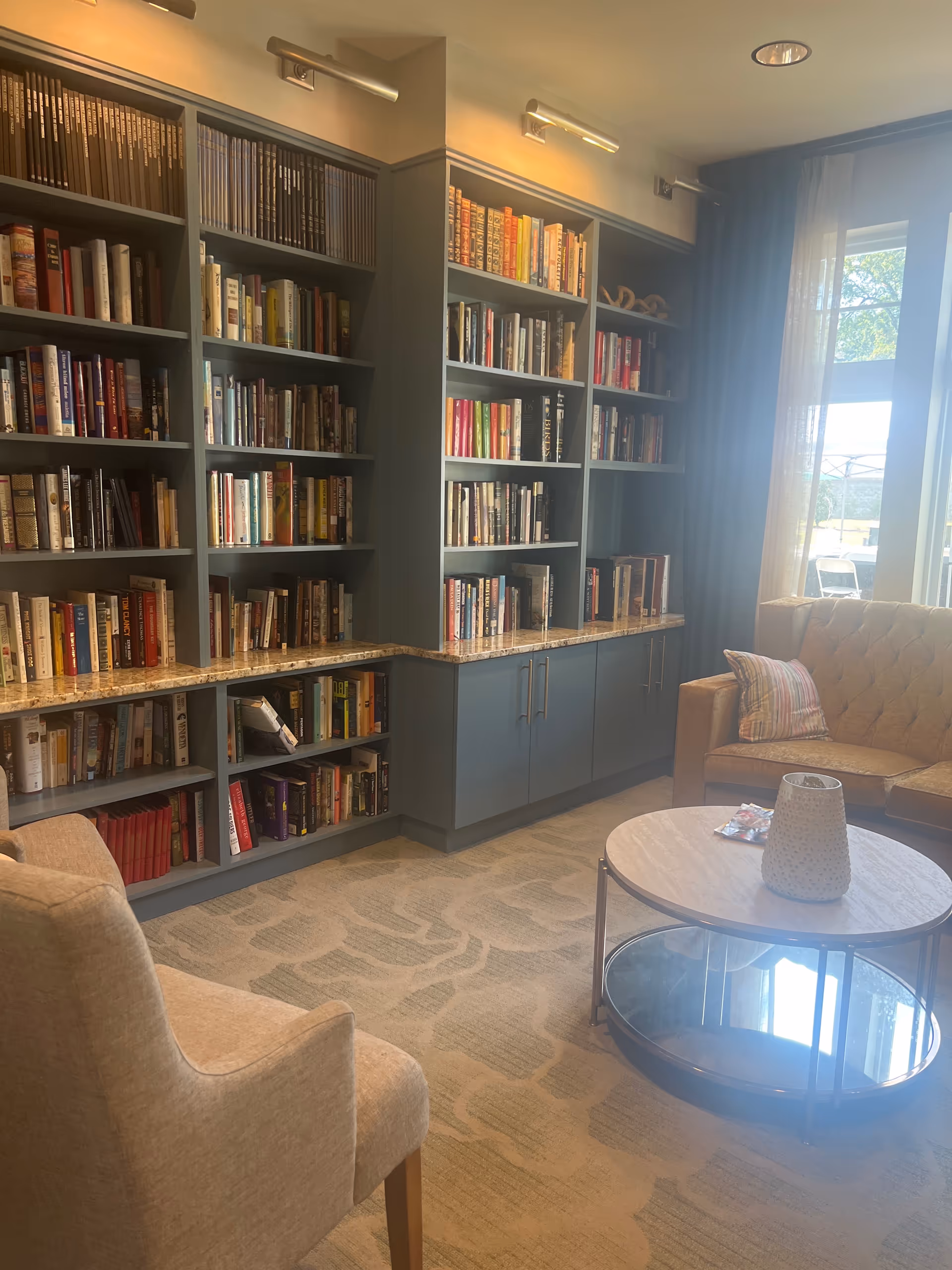 Interior library lounge with tall bookshelves, cabinets, and a sofa beside a bright window.
