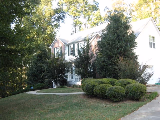 A two-story brick and white siding house surrounded by green bushes and trees with a concrete driveway and a small lawn in front.
