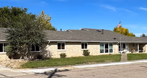 Single-story brick building with a gray shingled roof, several windows, and an American flag on a flagpole in front. The building is surrounded by a well-maintained lawn, a tree on the left side, and a clear blue sky above.
