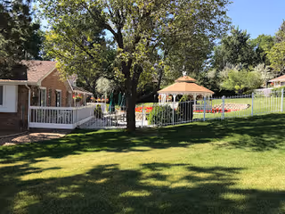 Outdoor area of a senior living facility with a large green lawn, a tree casting shadows, a white fence, a gazebo, and buildings in the background under a clear blue sky.