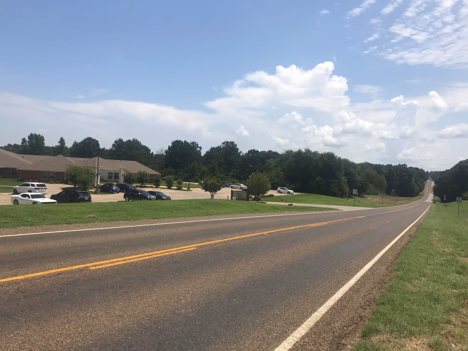 A long paved road stretches into the distance under a partly cloudy sky. On the left side of the road, there is a parking lot with several cars parked in front of a single-story building surrounded by trees and grass.