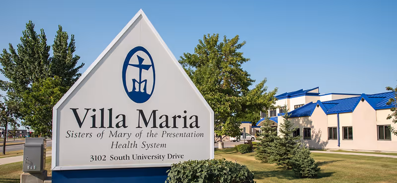 A large 'Villa Maria' entrance sign stands on a lawn in front of a beige building with blue roofs and trees.