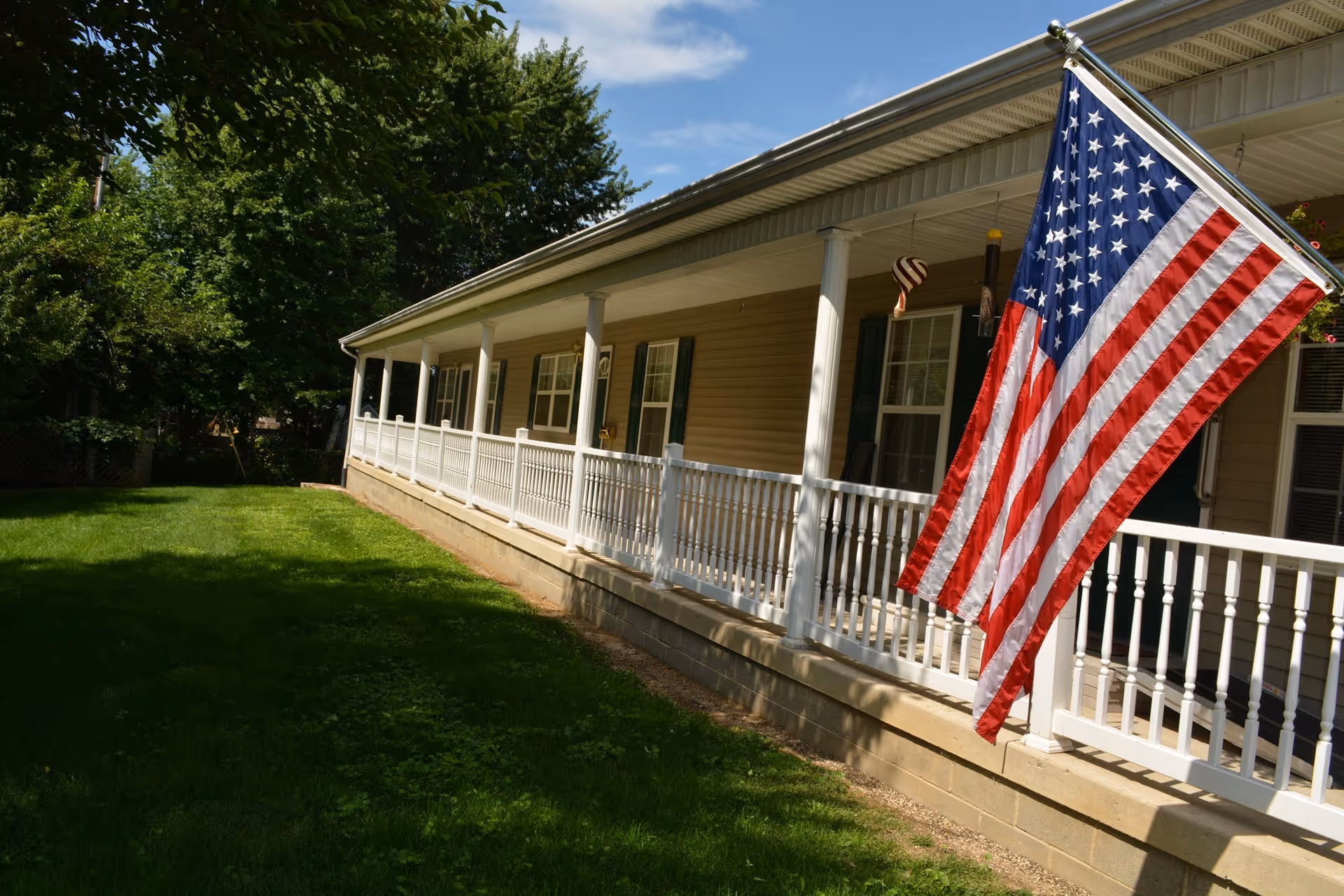 Long covered front porch of a single-story building with white railing, green lawn, and a large American flag.