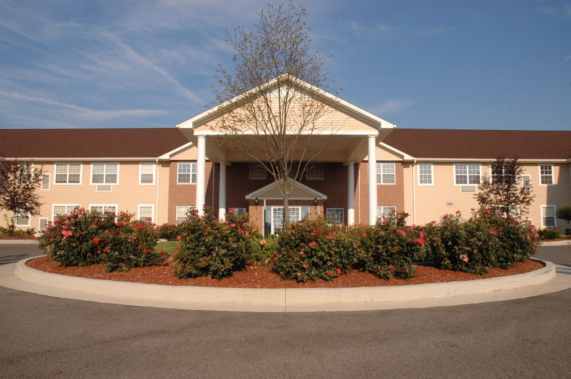 Front exterior view of a two-story senior living facility building with a covered entrance supported by white columns, surrounded by landscaped bushes and flowers under a clear blue sky.