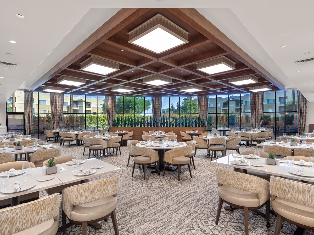 Bright, spacious dining room with multiple set tables and upholstered chairs beneath a coffered wooden ceiling and large windows.