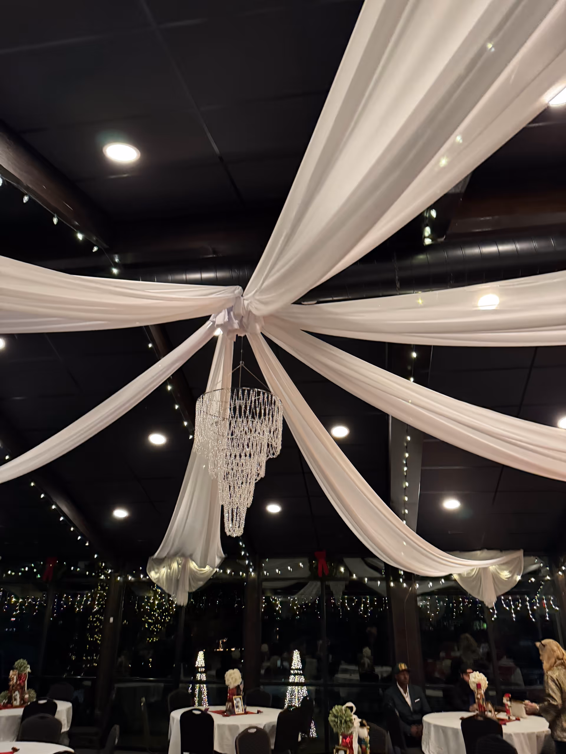 Elegant indoor dining hall with white drapery, chandeliers, and festive round tables.