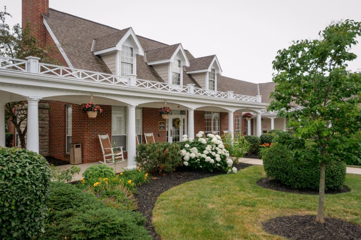 Brick building front with a covered white-column porch, rocking chairs, hanging flower baskets, and a landscaped lawn and shrubs.