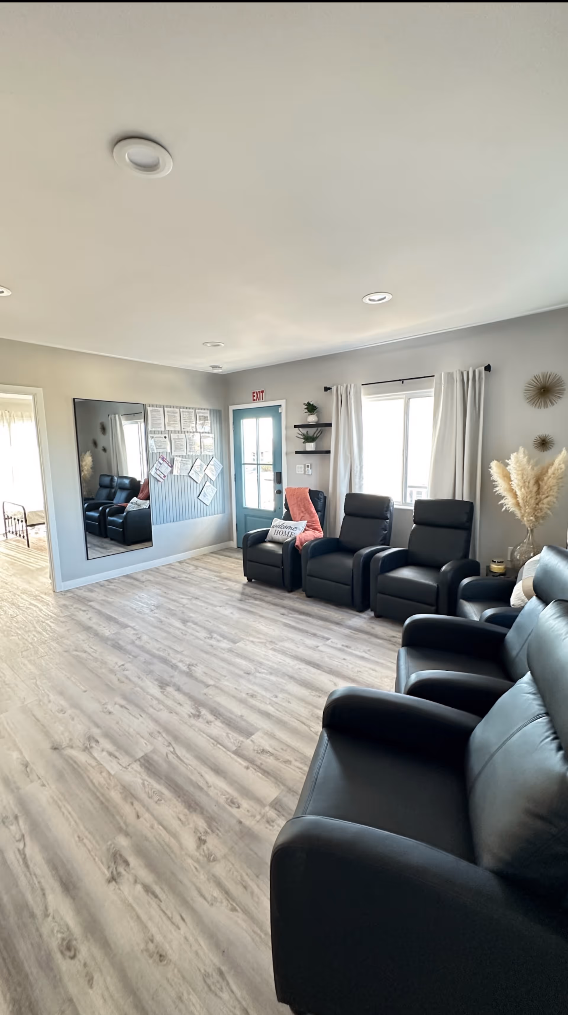 Bright communal living room with several black recliner chairs, a large wall mirror, a blue exit door and light wood flooring.