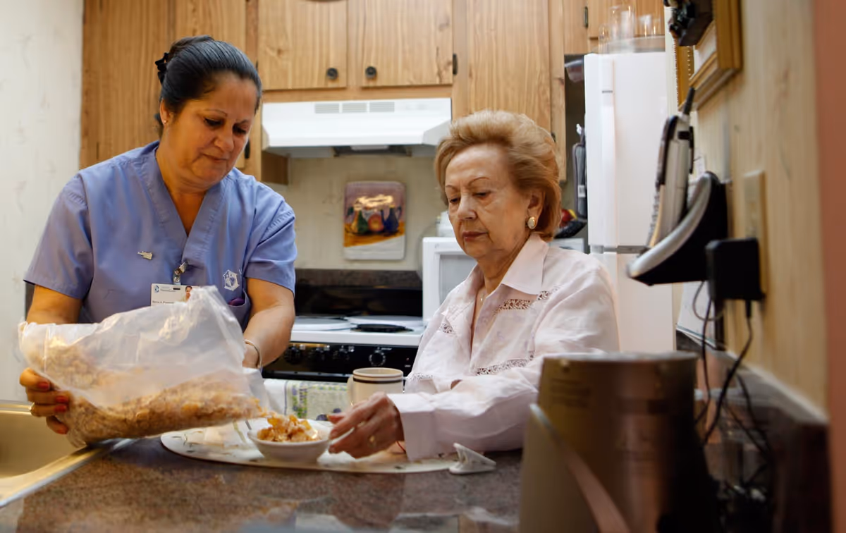 A caregiver in a blue uniform is helping an elderly woman in a white blouse by pouring cereal from a plastic bag into a bowl on a kitchen counter. The kitchen has wooden cabinets, a stove with a white hood, and various kitchen appliances.