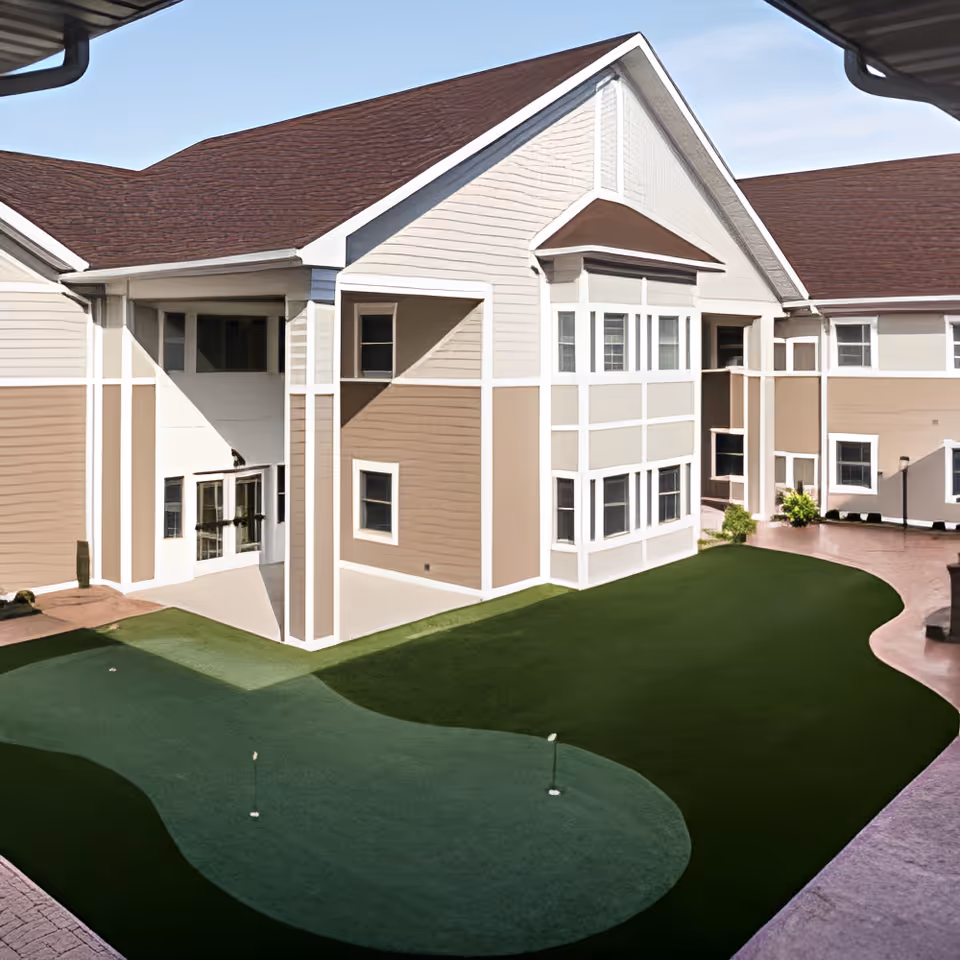 Outdoor courtyard area of Briarfield Place featuring a small artificial putting green surrounded by a paved walkway and beige multi-story building with white trim under a clear blue sky.