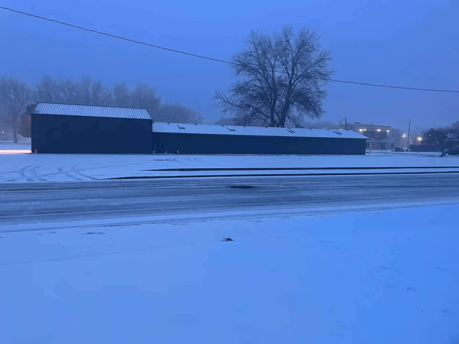 Snow-covered exterior of a long dark-blue building with a tall bare tree in the background.
