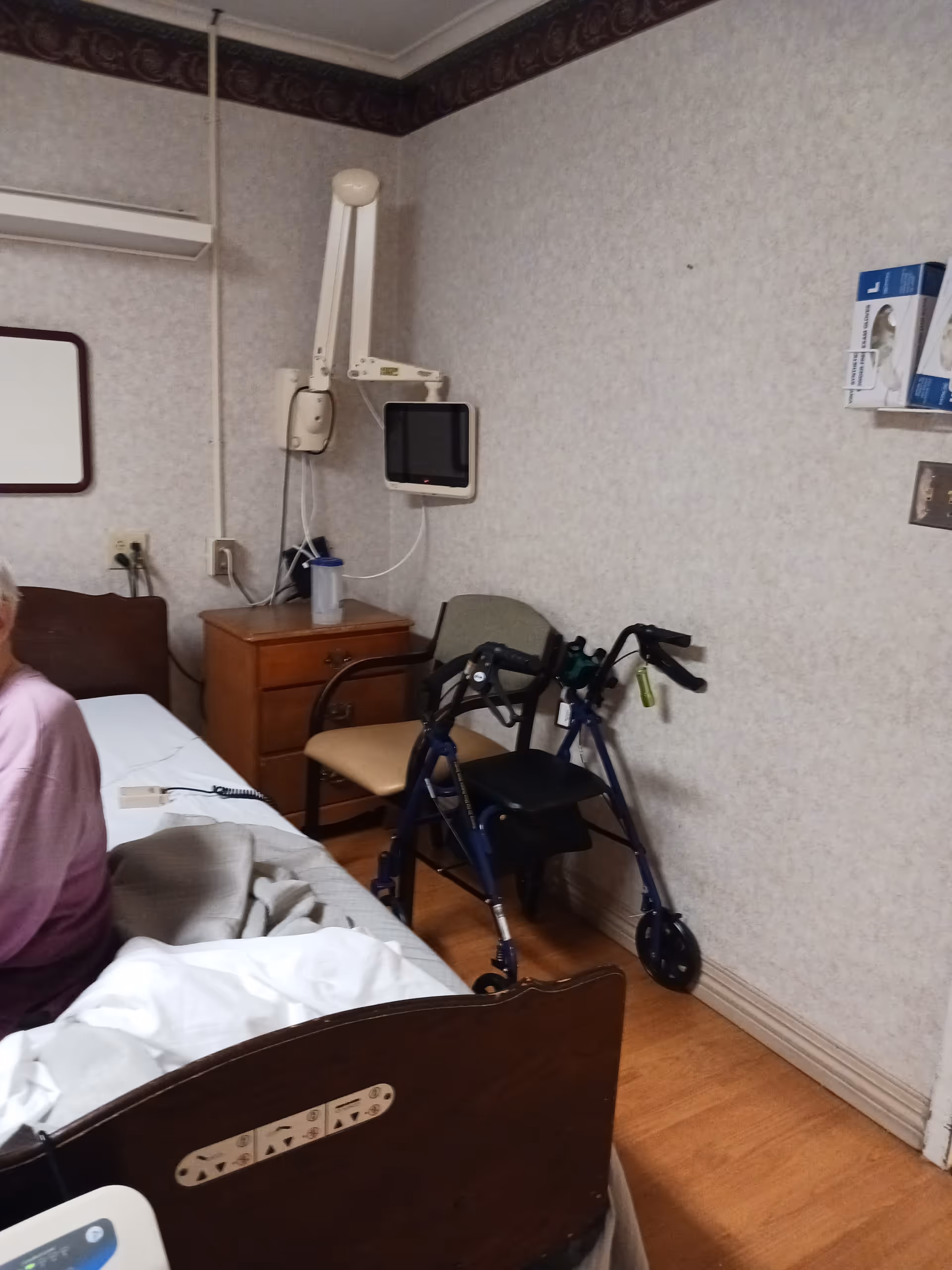 Patient room in a senior care facility with bed, walker, and monitor.