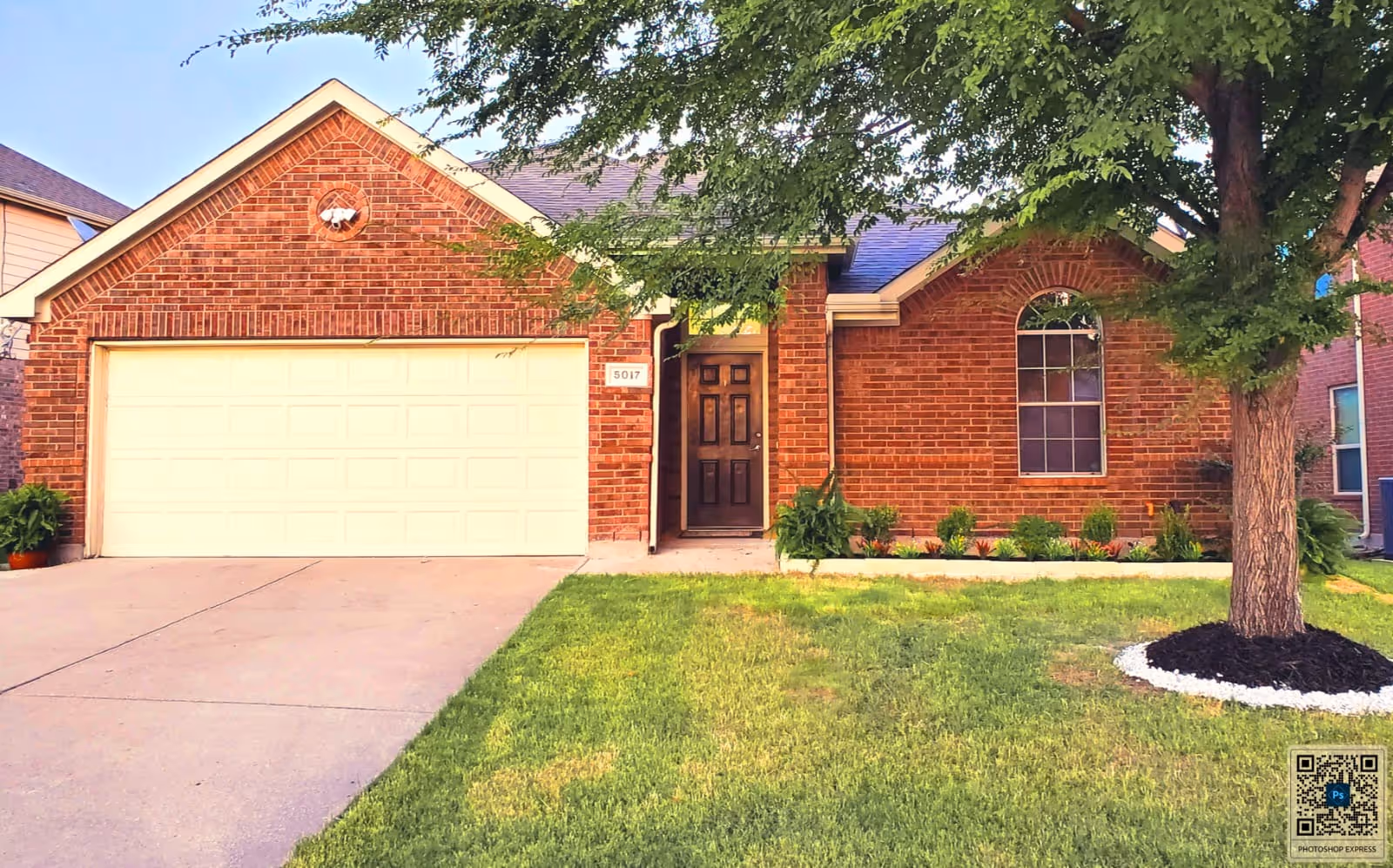 Brick residence with a two-car garage, front door, manicured lawn, and a tree.