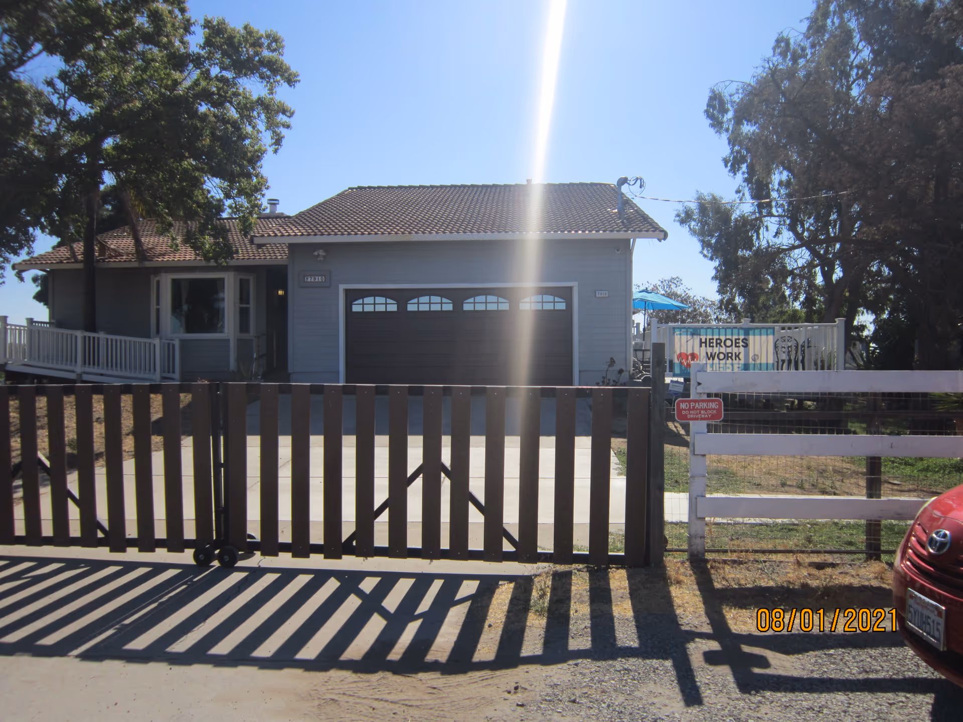 Front view of a single-story house with a closed garage and a wooden driveway gate, trees and a sign visible to the right.