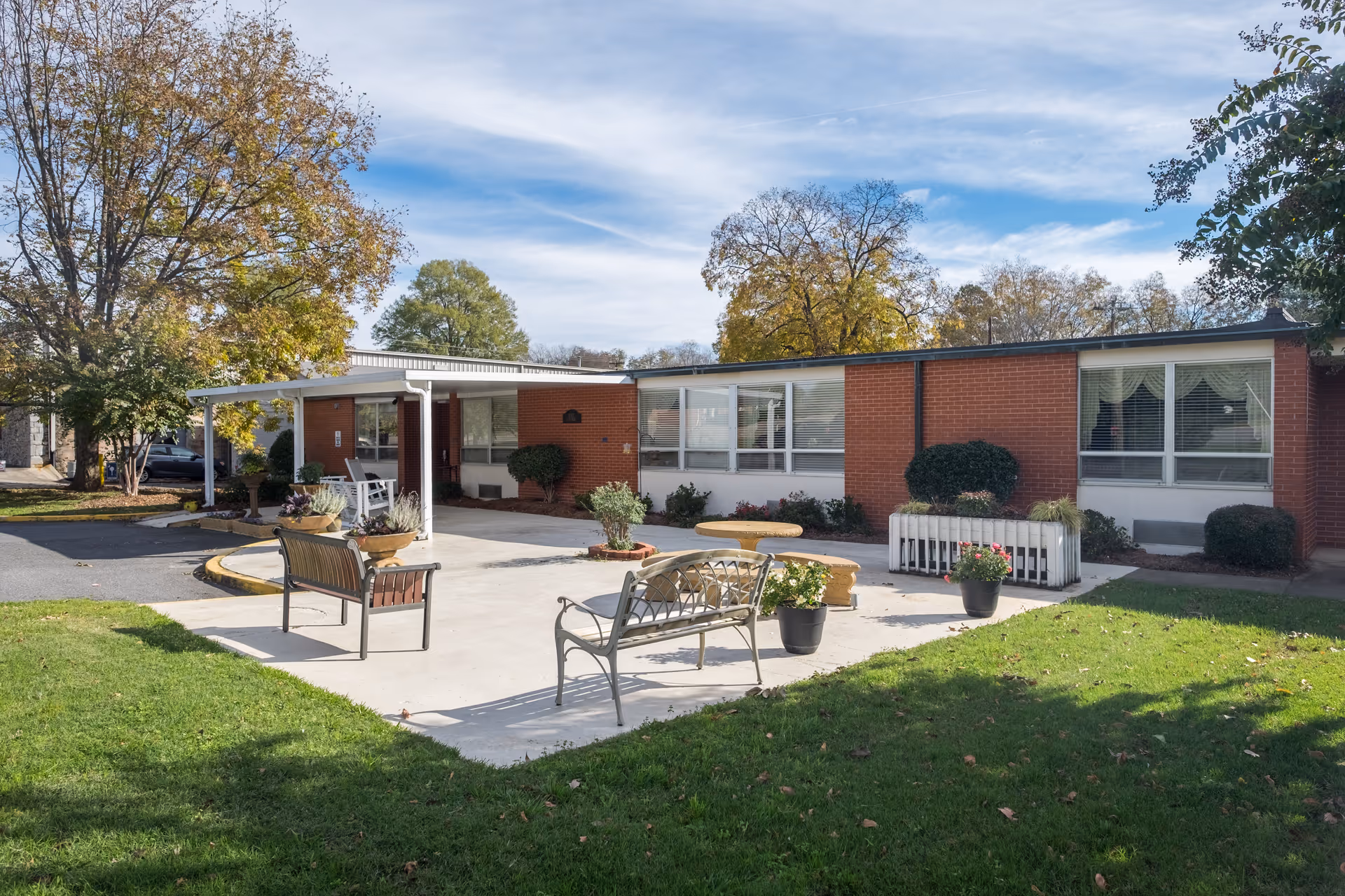 Single-story brick building front with a paved patio, benches, potted plants, and surrounding lawn and trees.