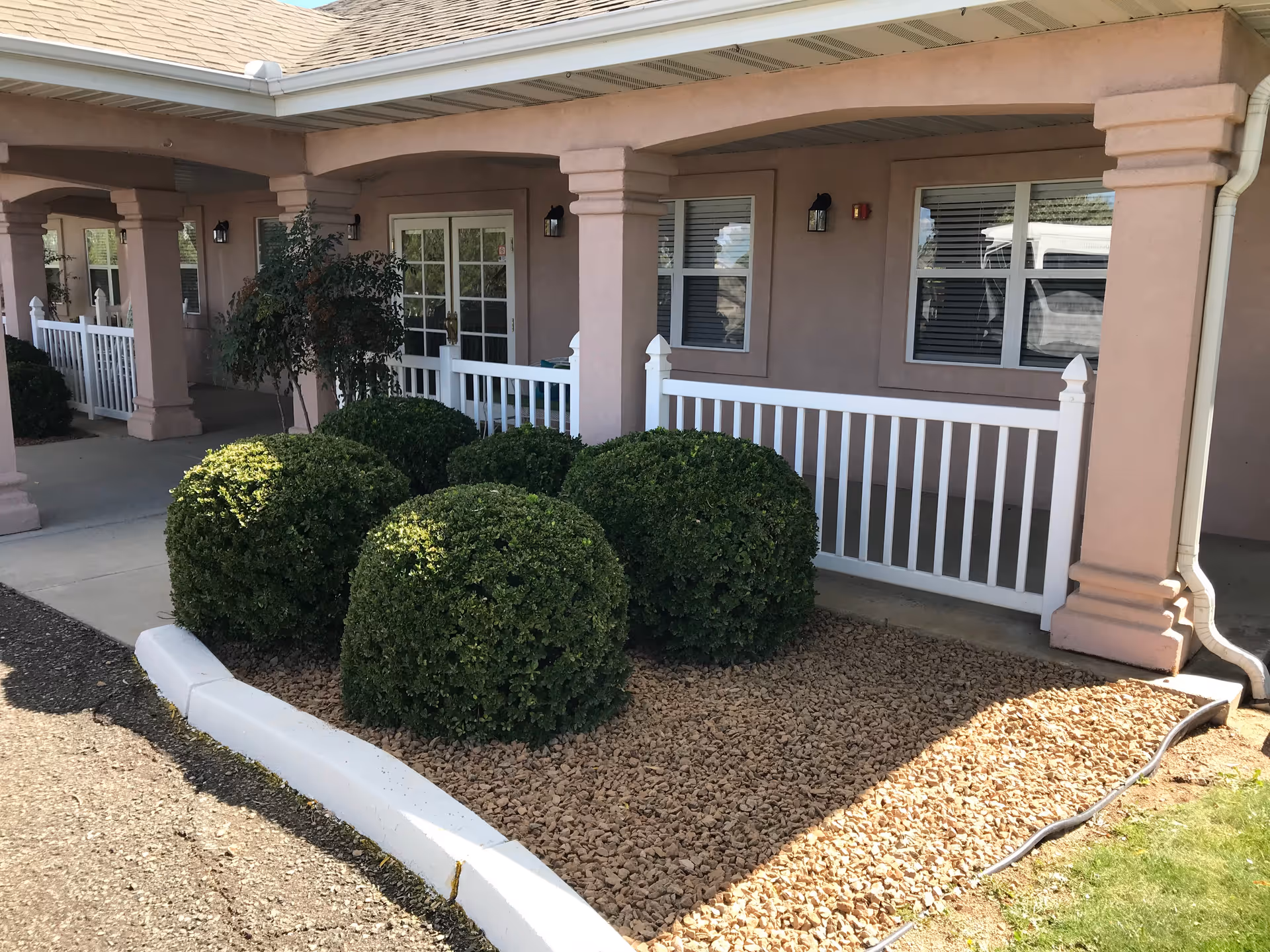 Front exterior of a senior living facility with pink walls, white railing, and trimmed bushes.