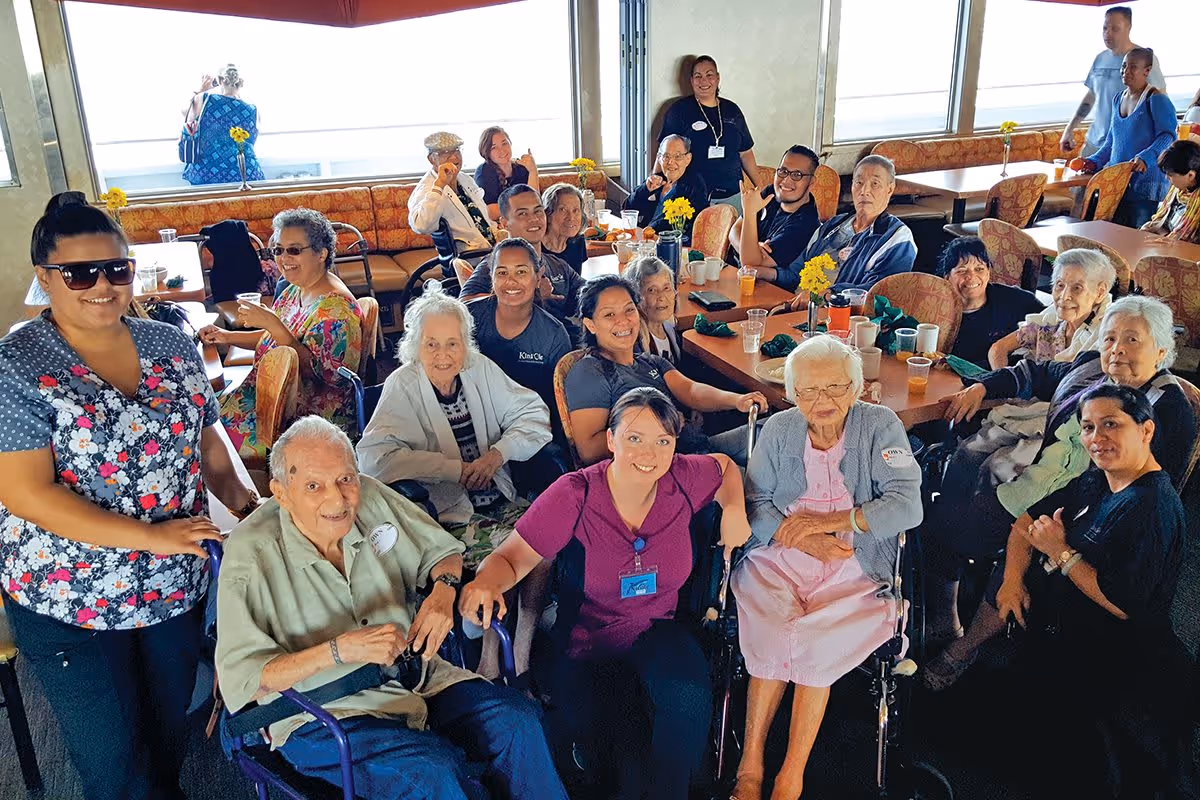 A group of seniors and staff members gathered around tables in a communal dining area.