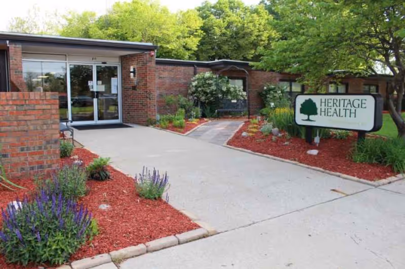 Front entrance of a low brick health facility with landscaped flowerbeds and a sign reading "HERITAGE HEALTH".