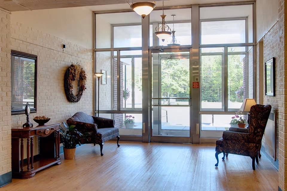 Bright lobby area with glass doors, two chairs, console table, wreath, and plants.