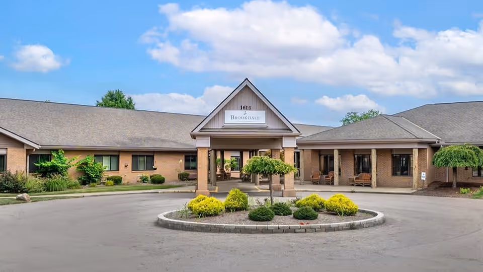 Front exterior view of Brookdale Mount Vernon facility showing a single-story brick building with a covered entrance, a circular driveway, and landscaped greenery under a partly cloudy sky.