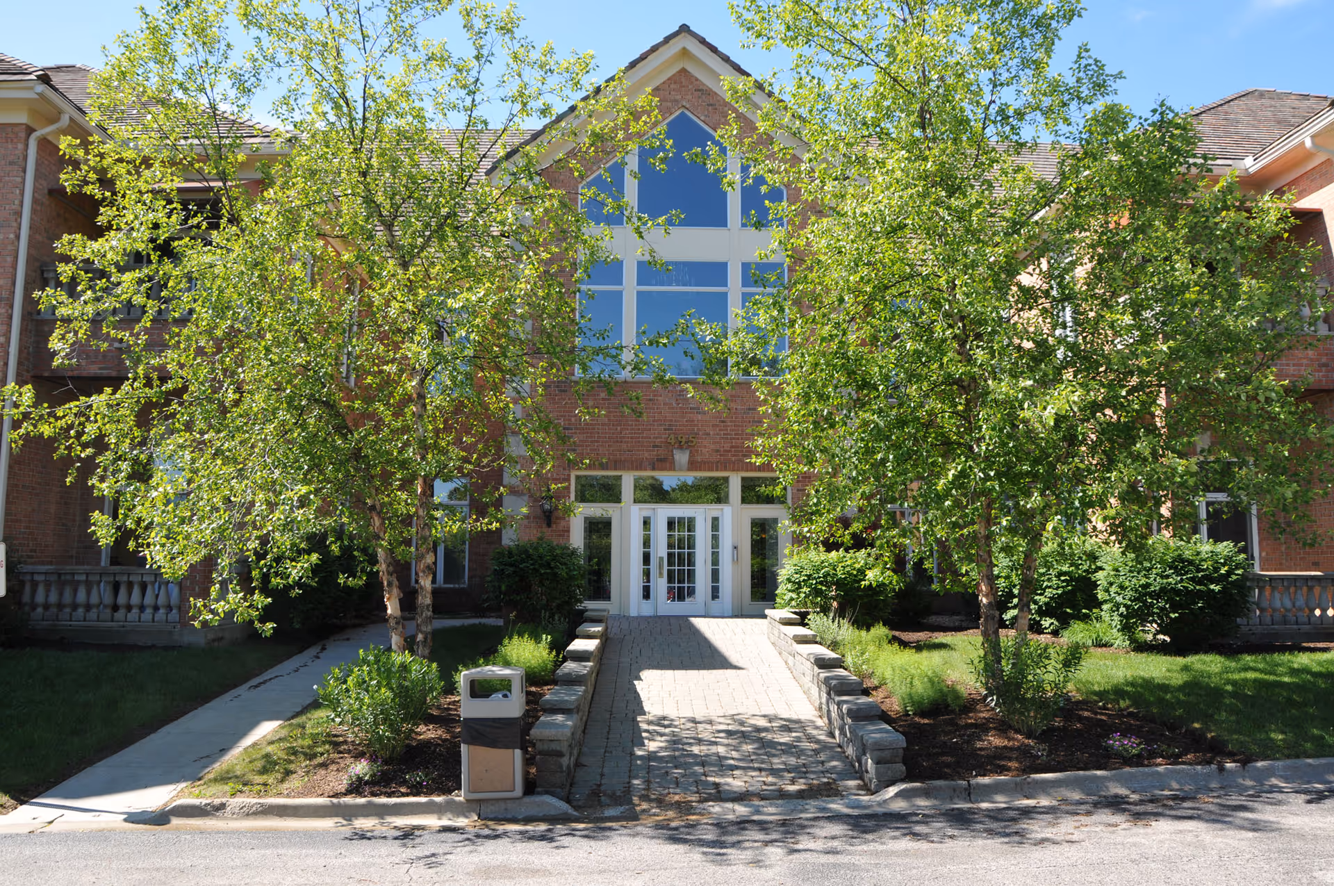 Brick senior living building entrance with glass doors, central walkway, and green trees.