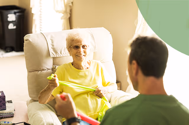 Elderly woman in yellow sits in a recliner, engaging with caregiver using resistance bands.
